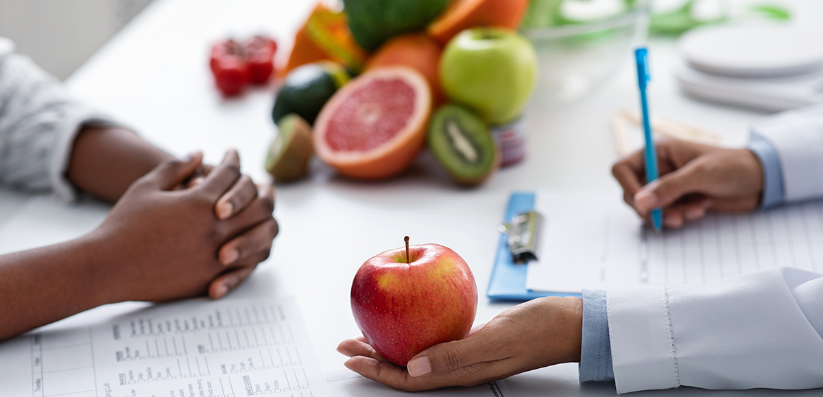Hands of a nutrition counselor with a fruit in one hand and writing with a pen with the other, and the hands of the person listening.