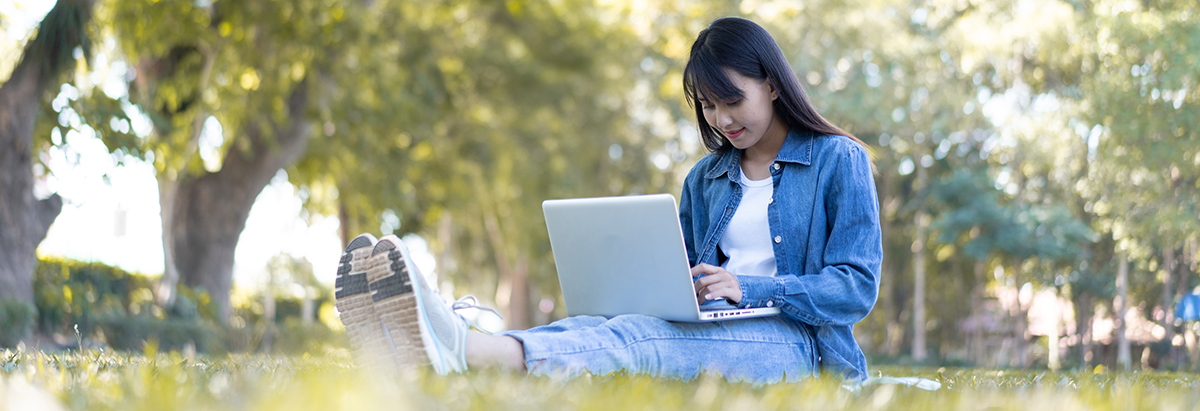 Asian student young woman with glasses sitting on the grass looking at her laptop.